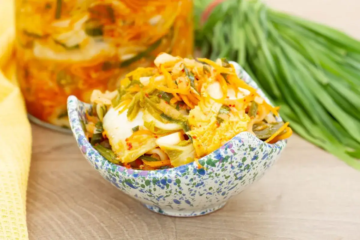 Kimchi co to? Pikantna, fermentowana kapusta pekińska z marchewką i szczypiorkiem w miseczce. W tle słoik z kimchi i pęczek zielonej cebulki.