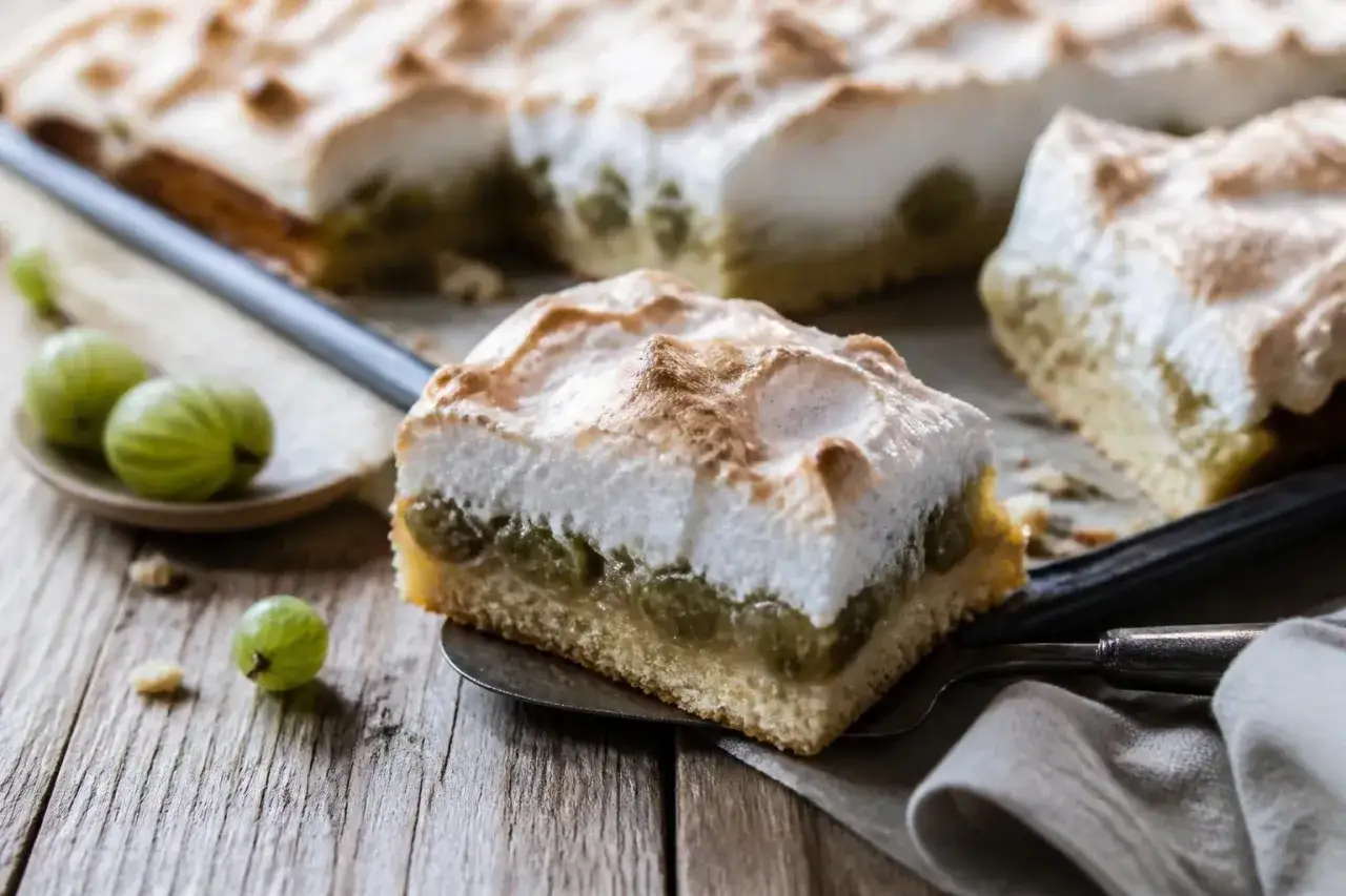 Ein St&uuml;ck vom besten Stachelbeerkuchen mit Baiserhaube auf einem Holztisch. Frische Stachelbeeren liegen daneben.