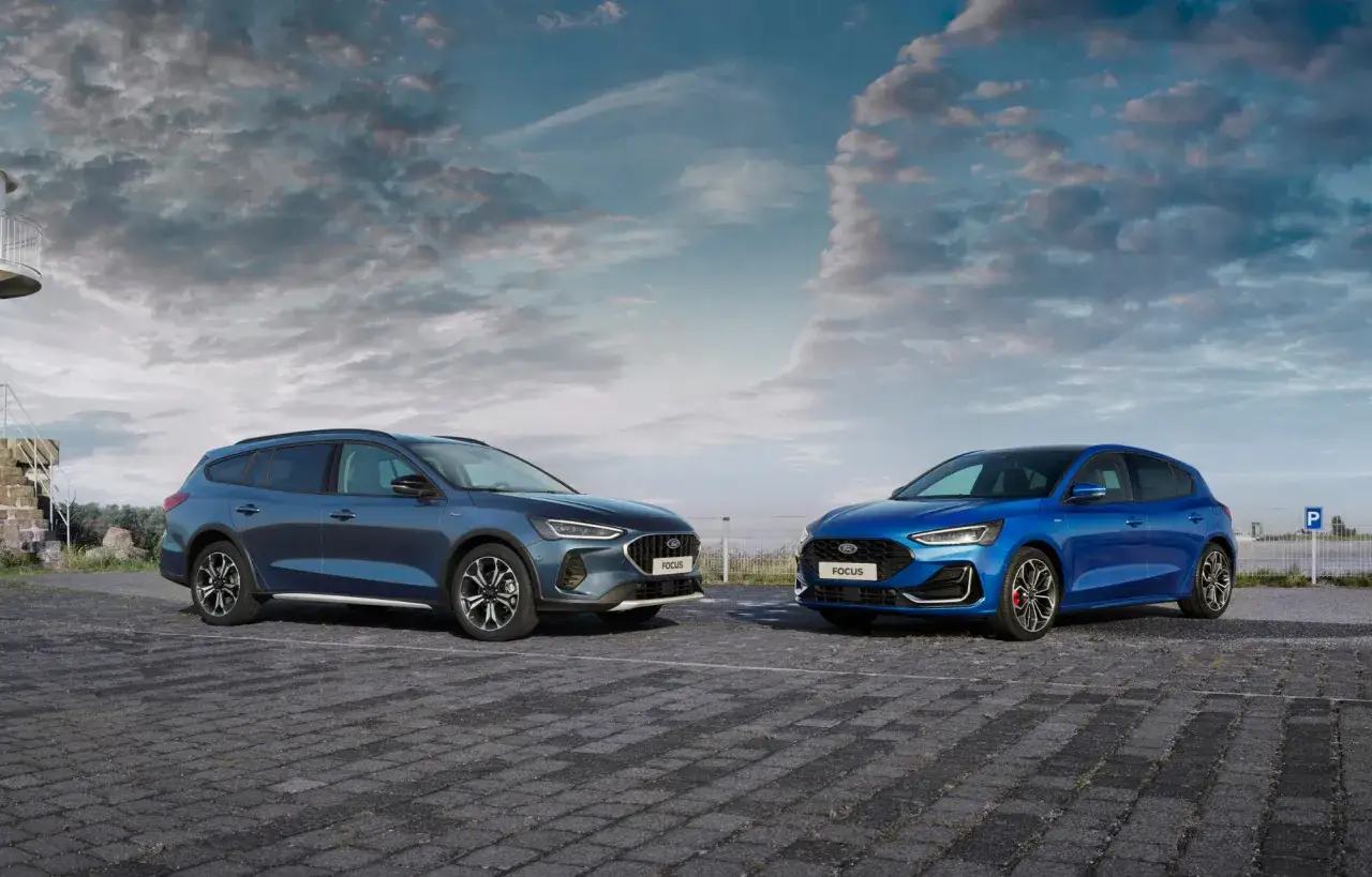Dwa modele Ford Focus 2025: kombi w kolorze ciemnoniebieskim i hatchback w kolorze jasnoniebieskim, stojące na placu pod pochmurnym niebem.