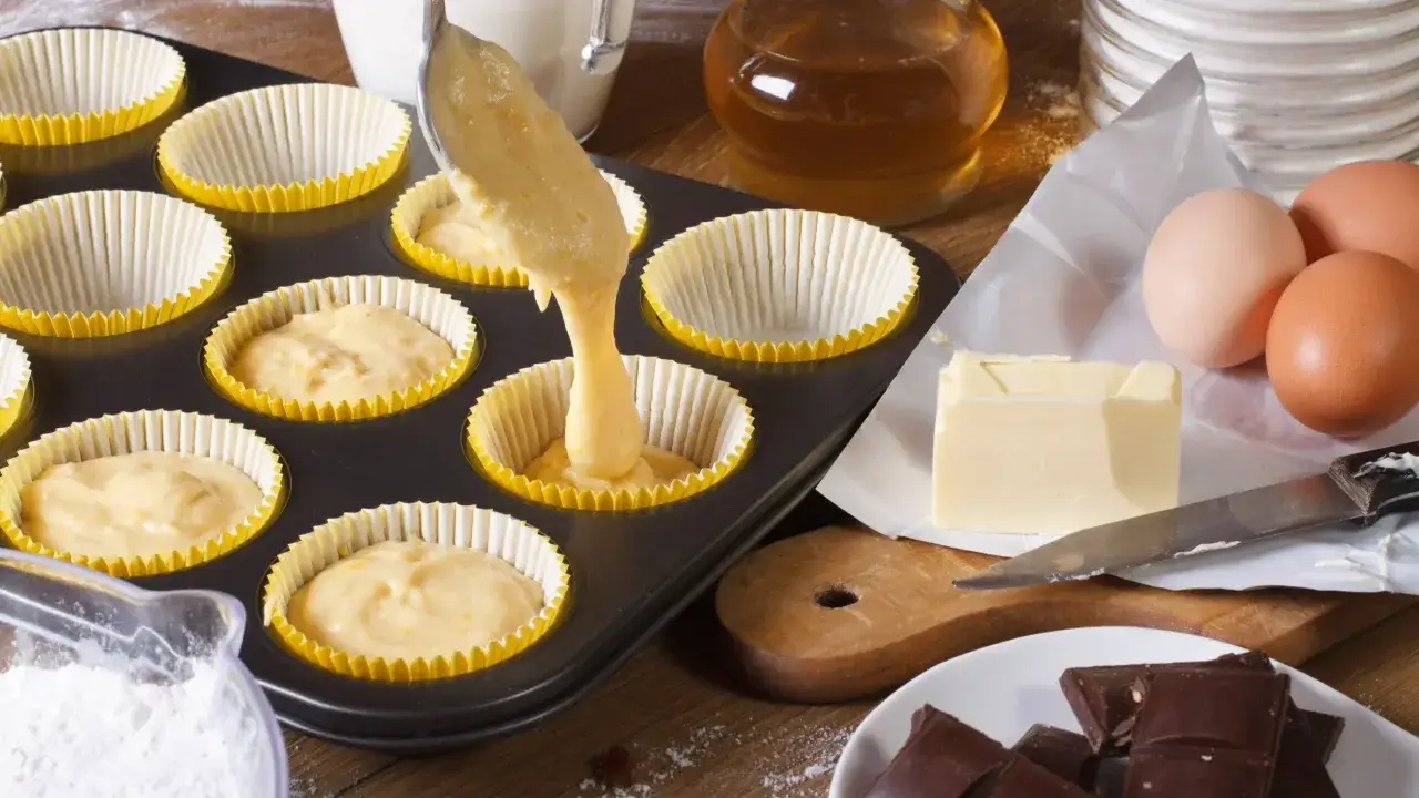Przygotowanie muffinek waniliowych z czekoladą. Ciasto wylewane do foremek, obok składniki: jajka, masło, czekolada.