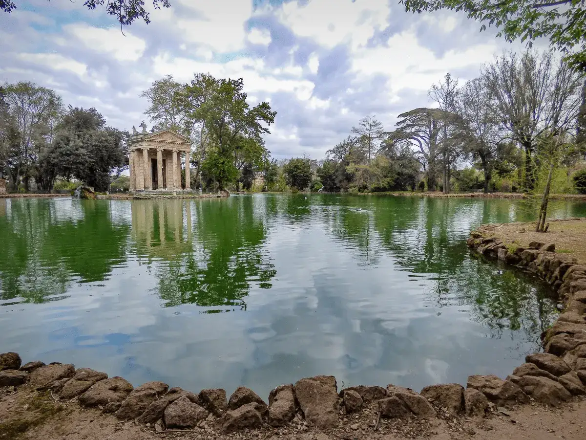 Tempio di Esculapio sull'acqua, riflesso nel lago di Villa Borghese. Alberi verdi e cielo nuvoloso.