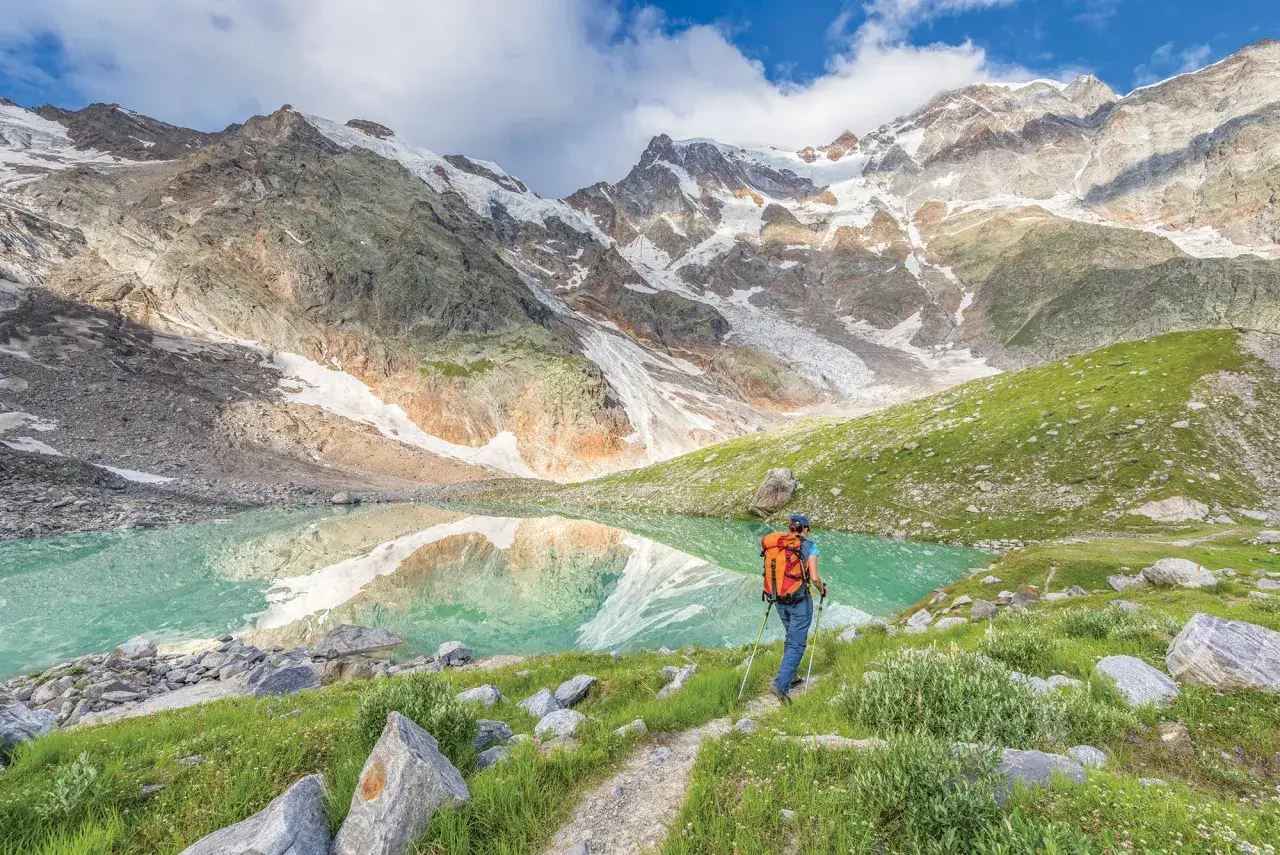 Trekking w Alpach: turystka z plecakiem idzie ścieżką nad turkusowym jeziorem, w tle skaliste szczyty z lodowcami.