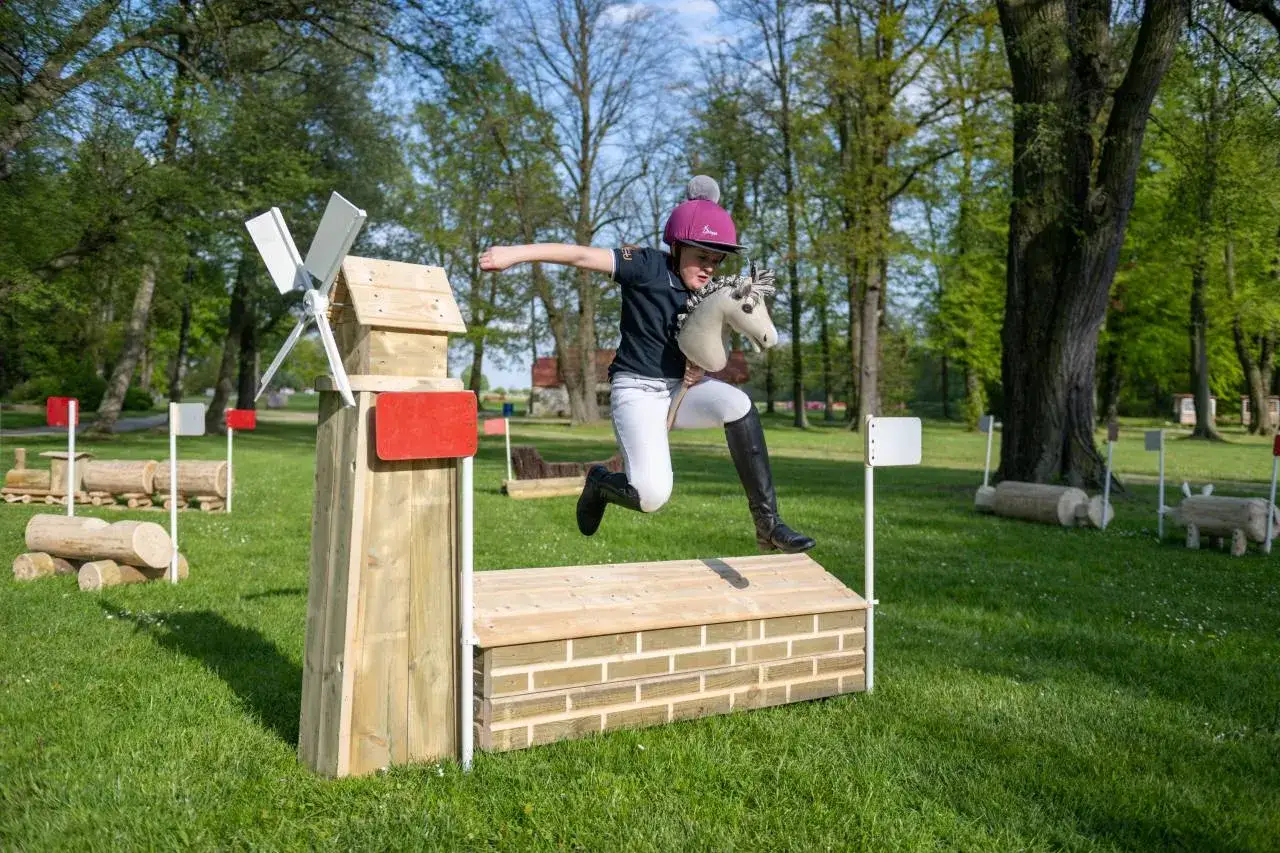Dziewczynka w kasku skacze przez przeszkodę z hobby horse. Czy hobby horse to sport? Tak, to aktywność wymagająca zręczności i koordynacji.