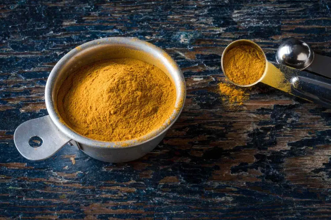 Une cuillère doseuse remplie d'épice colombo, d'une belle couleur jaune orangé, repose sur une surface en bois sombre.