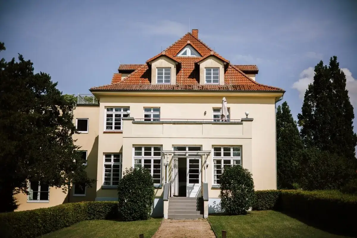 Ein gelbes Haus mit roten Ziegeldach und vielen Fenstern, umgeben von grünen Bäumen und Büschen. Ein **gut gremmelin**er Ort für einen entspannten Tag.