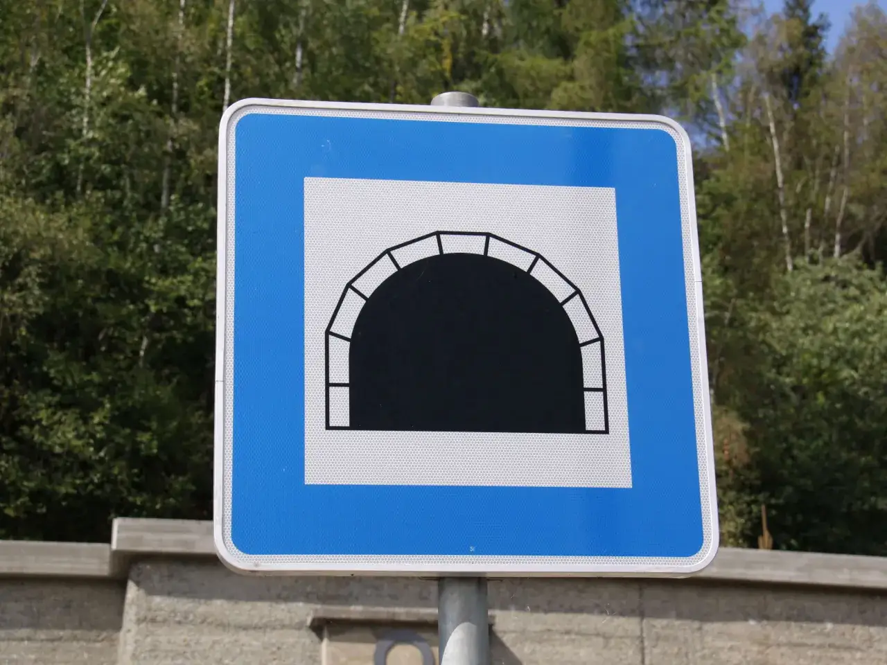 Blaues Verkehrsschild mit einem wei&szlig;en Quadrat, das einen schwarzen Tunnel zeigt. Achtung, bald kommt ein Tunnel!