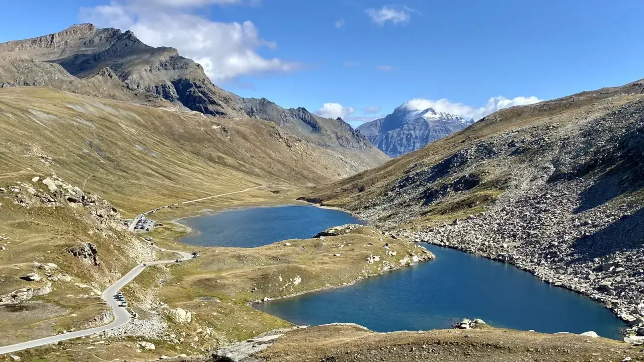 Due laghi alpini blu intenso riflettono il cielo azzurro al Col del Nivolet, con una strada tortuosa che serpeggia tra montagne rocciose e pendii erbosi.