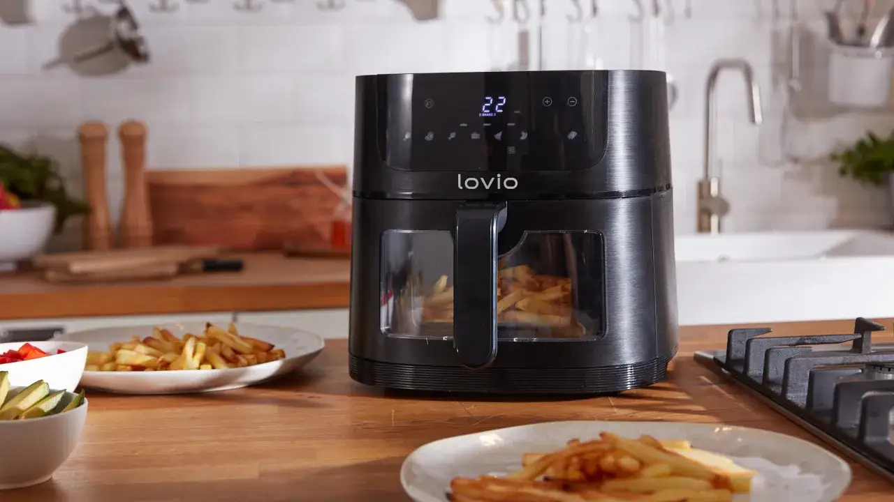 Czarny, nowoczesny air fryer LoviO z okienkiem, w którym widać złociste frytki. Obok talerze z gotowymi frytkami i miseczki z warzywami. To najlepszy air fryer do szybkiego przygotowania chrupiących przekąsek.