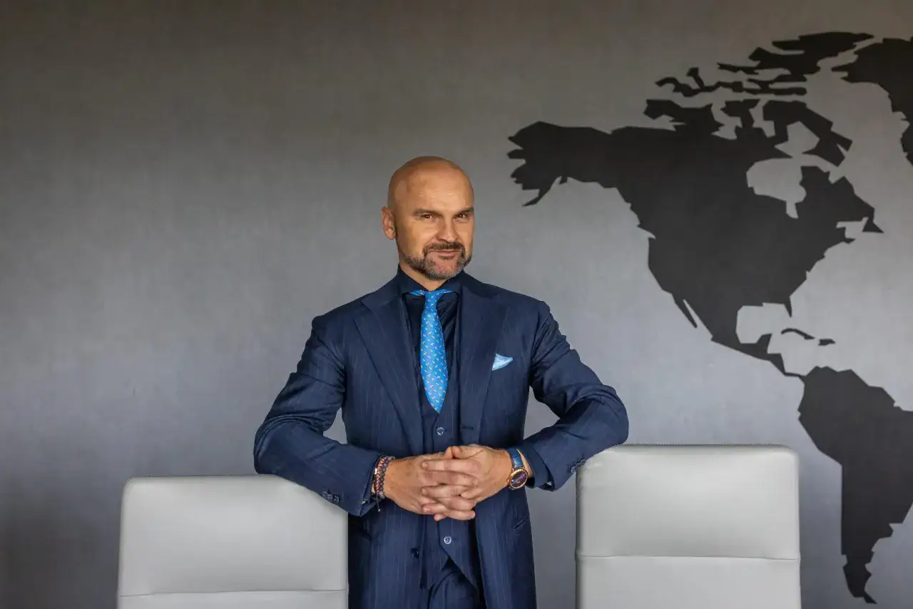 Rafał Brzoska, w eleganckim garniturze, z rękami złożonymi na piersi, na tle mapy świata. Jego majątek to symbol globalnych ambicji.
