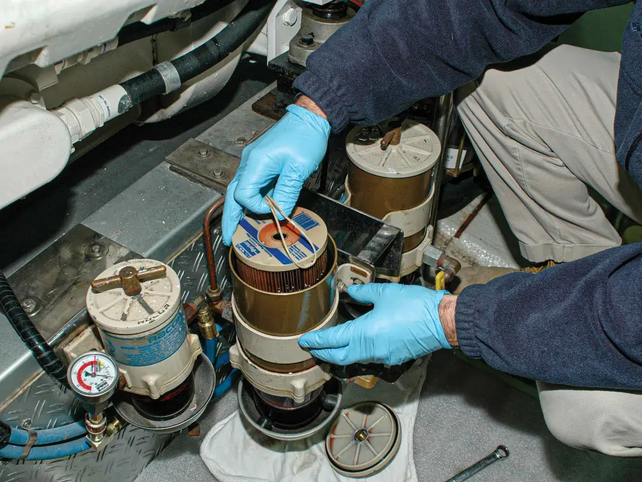 Un mécanicien en gants bleus change le filtre à carburant d'un moteur. On voit ici où se trouve le filtre à carburant, prêt à être remplacé.