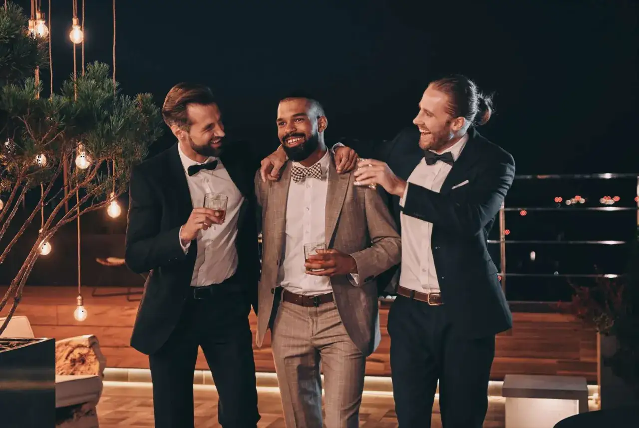 Three men in stylish suits and bow ties, holding drinks, offer inspiration for what to wear to a wedding men.