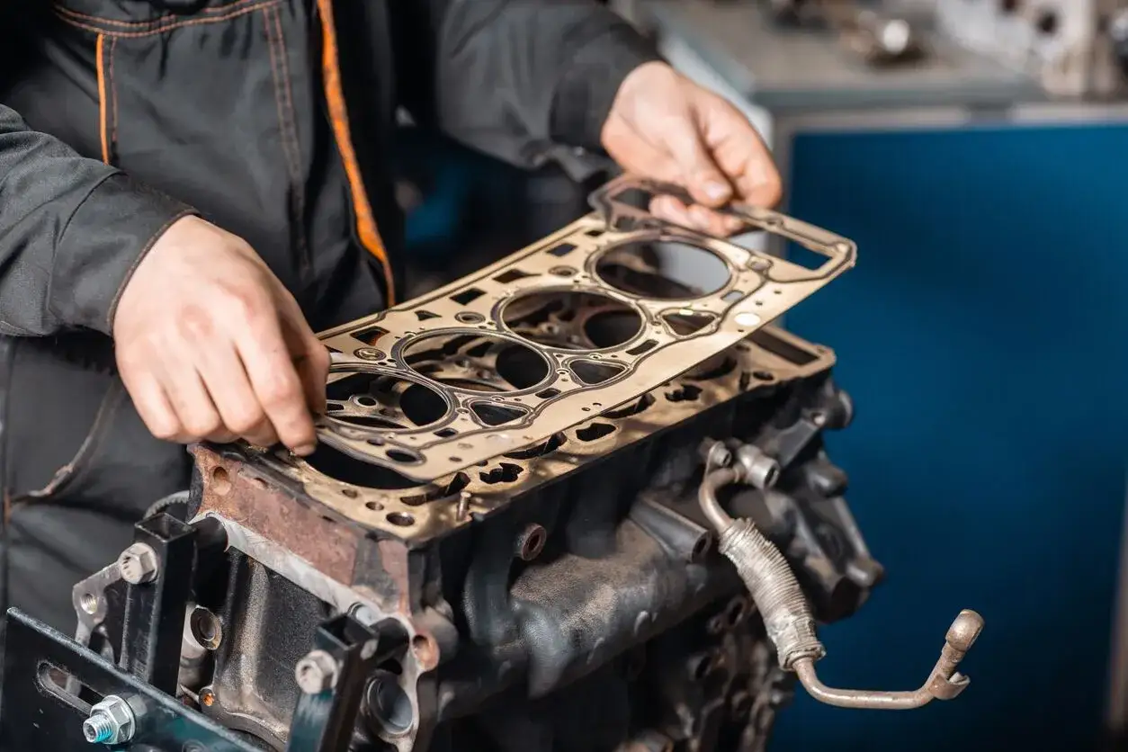 Mechaniker h&auml;lt Zylinderkopfdichtung &uuml;ber Motorblock. Ein Motorschaden kann hohe Kosten verursachen, aber eine Reparatur ist oft m&ouml;glich.