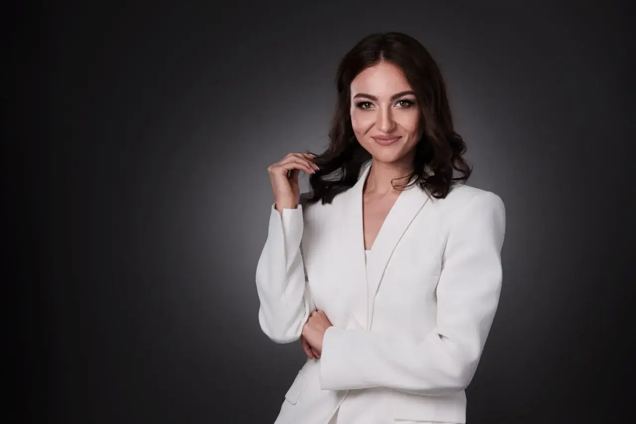 Profesjonalne cv photo: uśmiechnięta kobieta w białej marynarce, z dłonią przy włosach, na ciemnym tle.