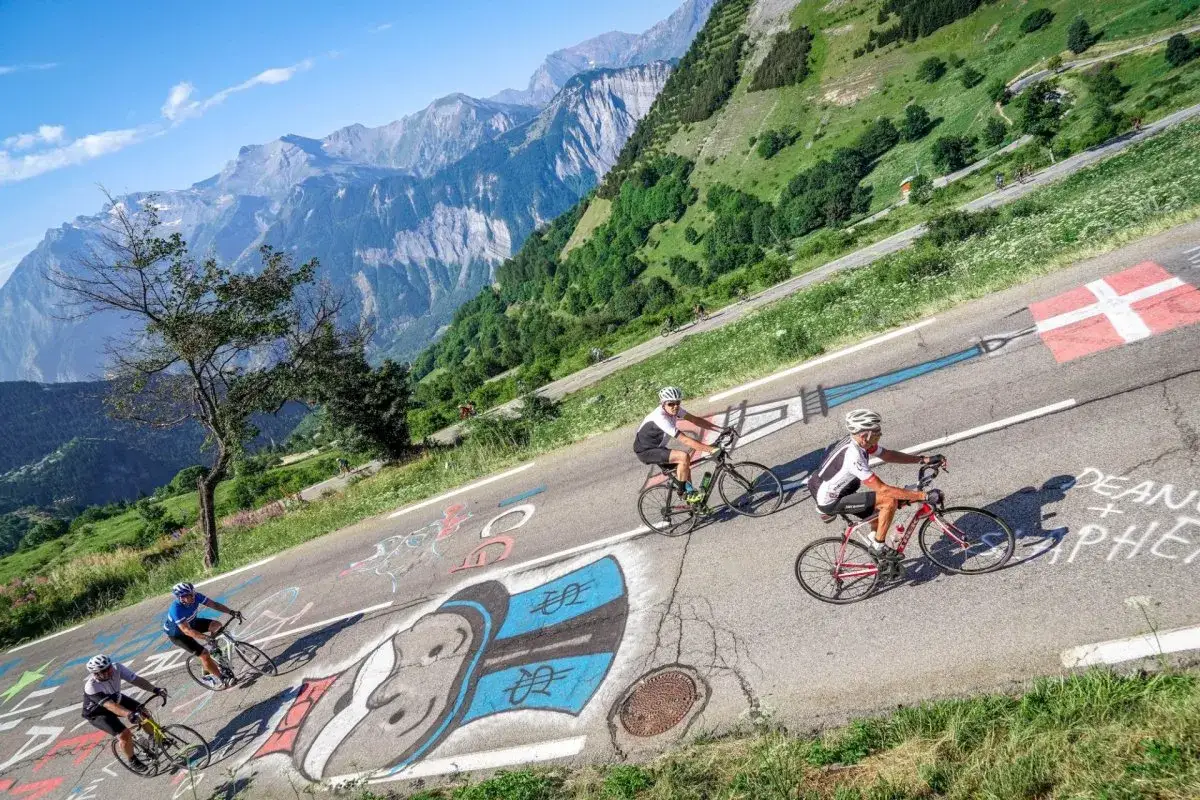Cyclists ascend the iconic Alpe d'Huez Tour de France climb, with graffiti and a Swiss flag marking the challenging route through the stunning French Alps.