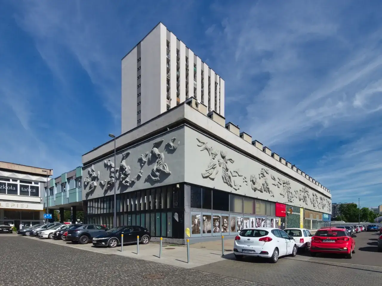 Fasada budynku z płaskorzeźbami, parking i wystawa na zewnątrz. To galeria sztuki Katowice.