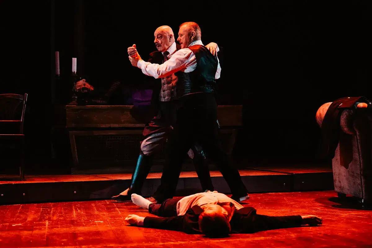 Dwóch mężczyzn tańczy tango, podczas gdy trzeci leży na scenie. Scena oświetlona jest na czerwono, tworząc atmosferę groteski w tangu.