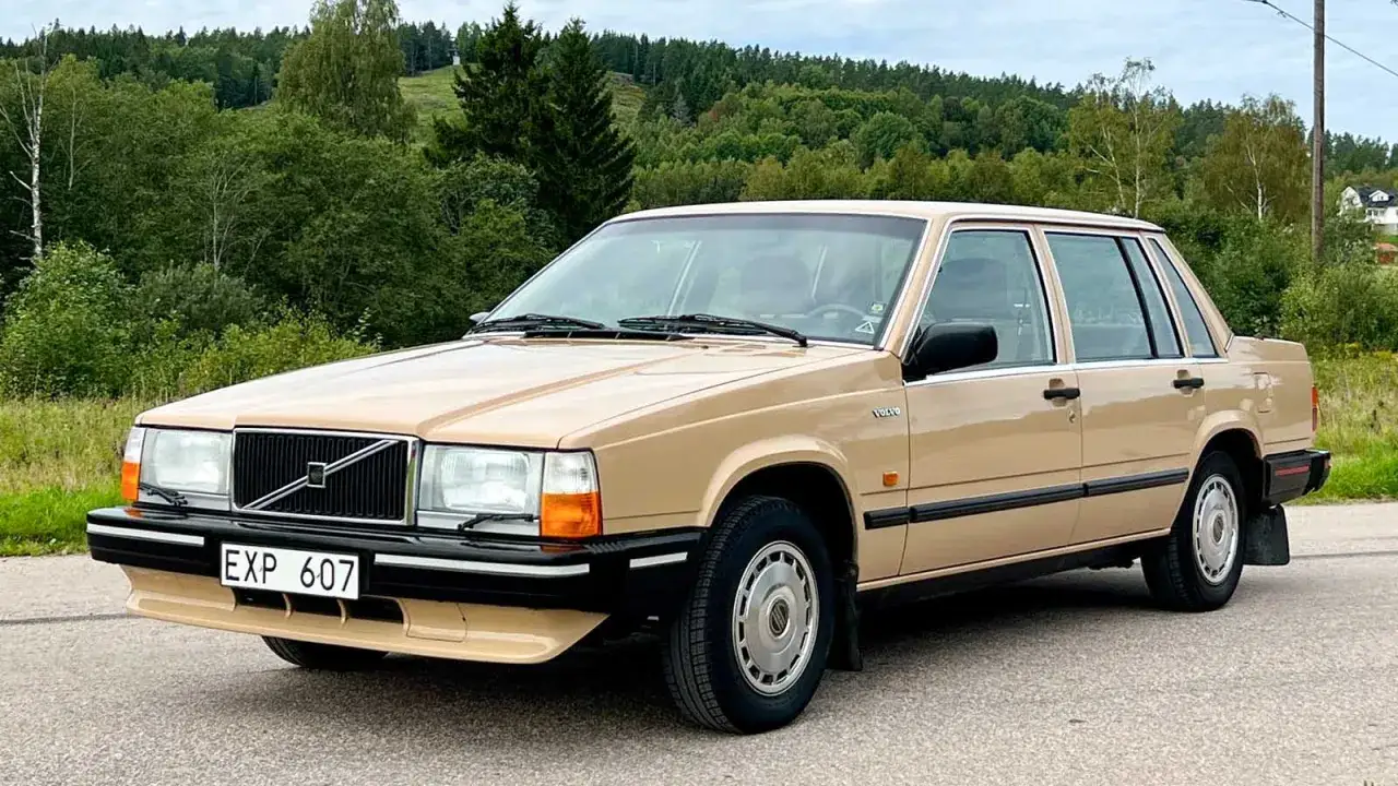 En beige Volvo 740 fr&aring;n 80-talet st&aring;r parkerad p&aring; en v&auml;g med en lummig skog i bakgrunden.