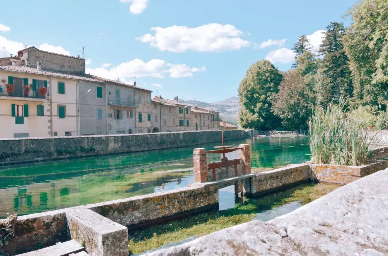 Un canale d'acqua verde smeraldo scorre tra case antiche e vegetazione lussureggiante, con il Monte Amiata sullo sfondo.