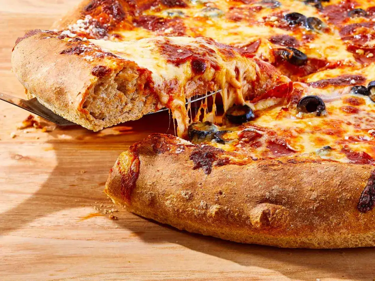 Ein Stück vollkorn pizza mit geschmolzenem Käse wird herausgehoben. Darauf sind Salami und Oliven zu sehen.