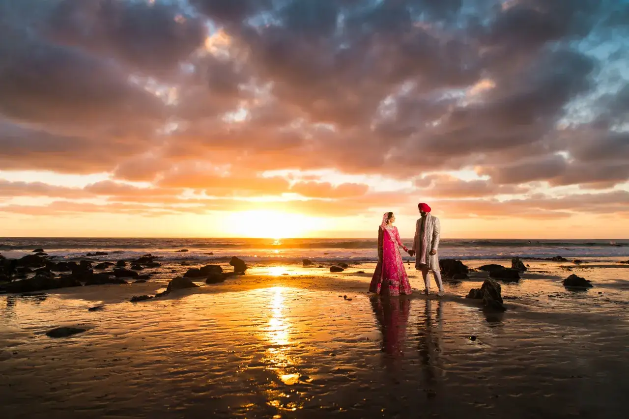 Un couple en tenue traditionnelle indienne se tient la main sur une plage au coucher du soleil. Ces photos de plage capturent un moment magique.
