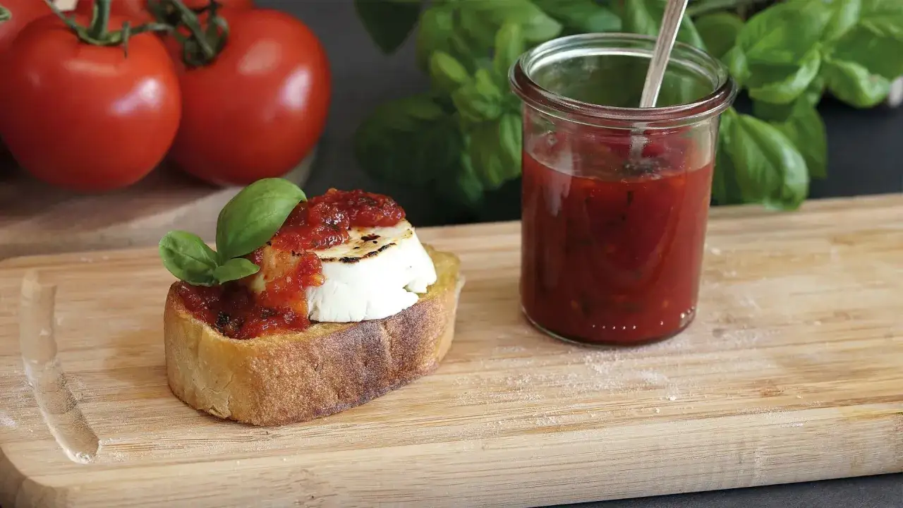 Bruschetta mit Ziegenk&auml;se und Tomatenmarmelade, inspiriert von einem italienischen Tomatenmarmelade Rezept. Frische Tomaten und Basilikum im Hintergrund.