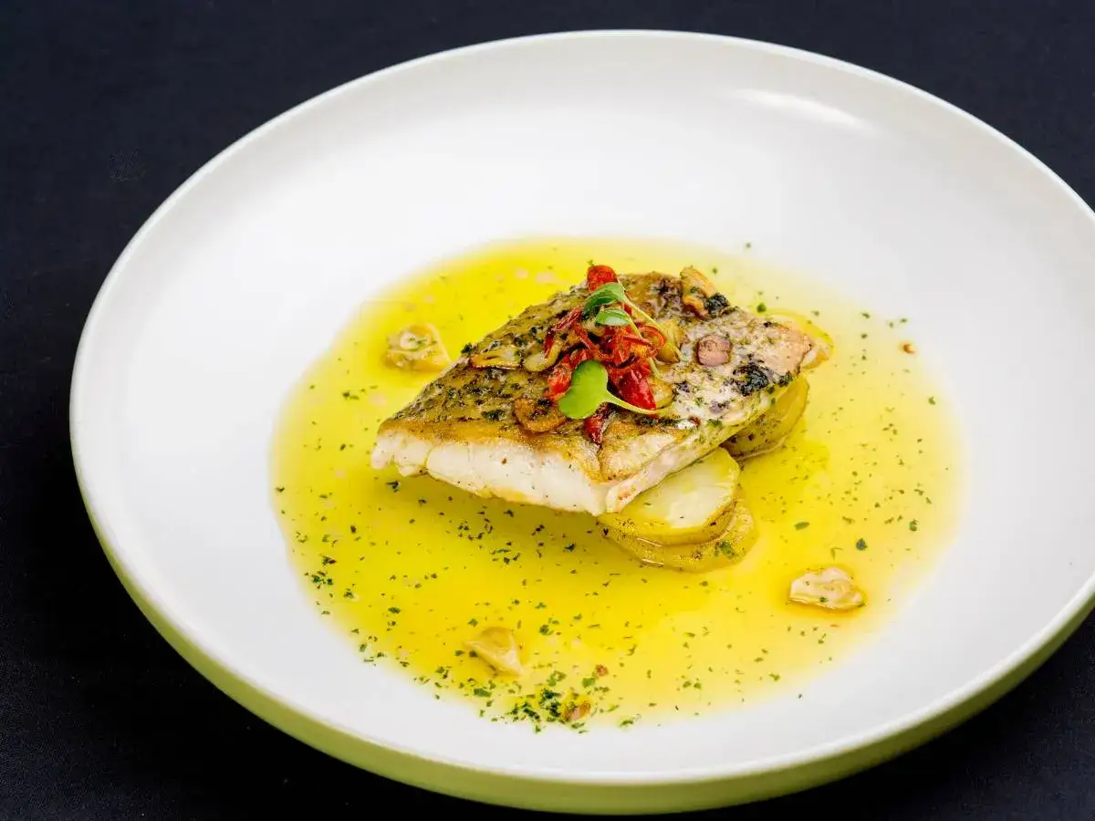 Filet ryby na plastrach ziemniaków, polany aromatycznym **sosem maślano cytrynowym**, ozdobiony ziołami i suszonymi pomidorami.