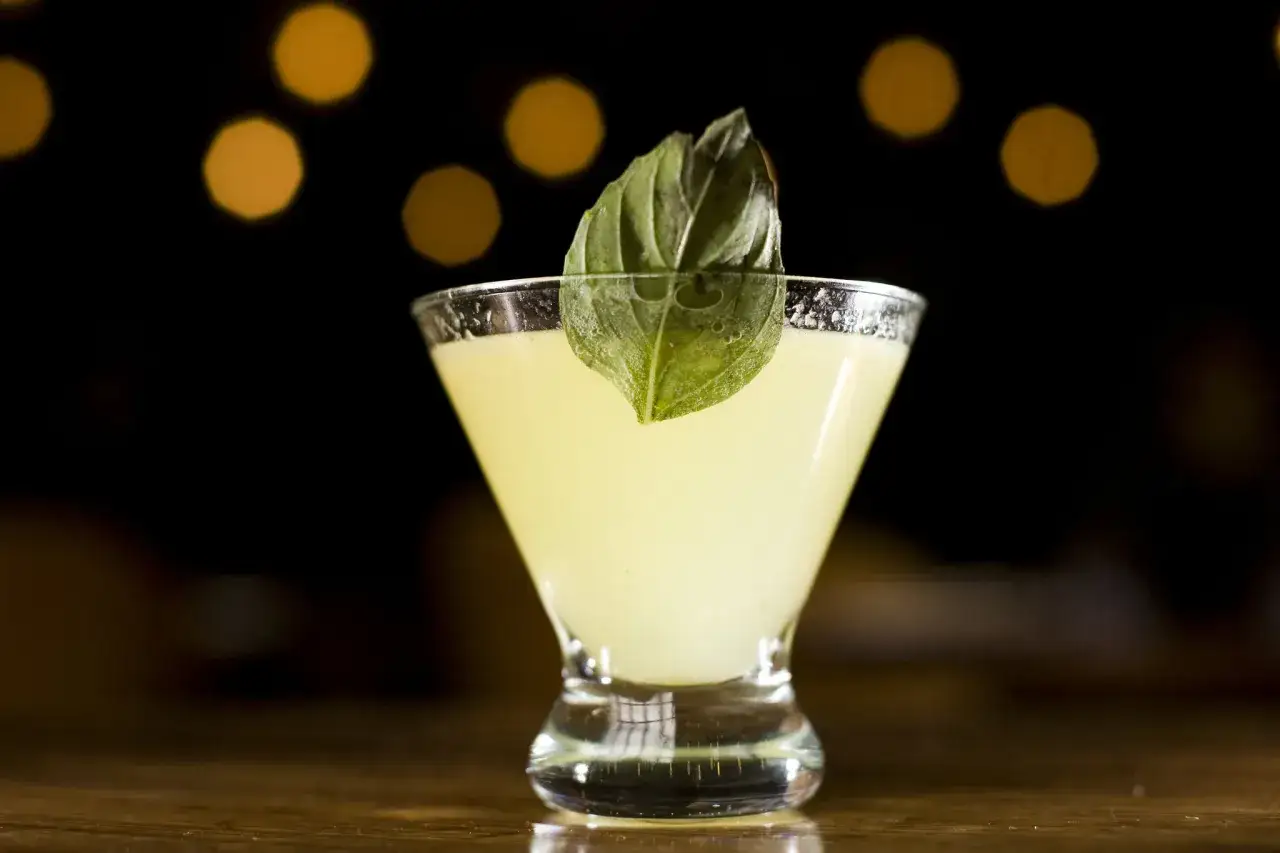 En ljusgul drink i ett martiniglas, garnerad med ett grönt blad. En perfekt thai basil drink för en sommarkväll.