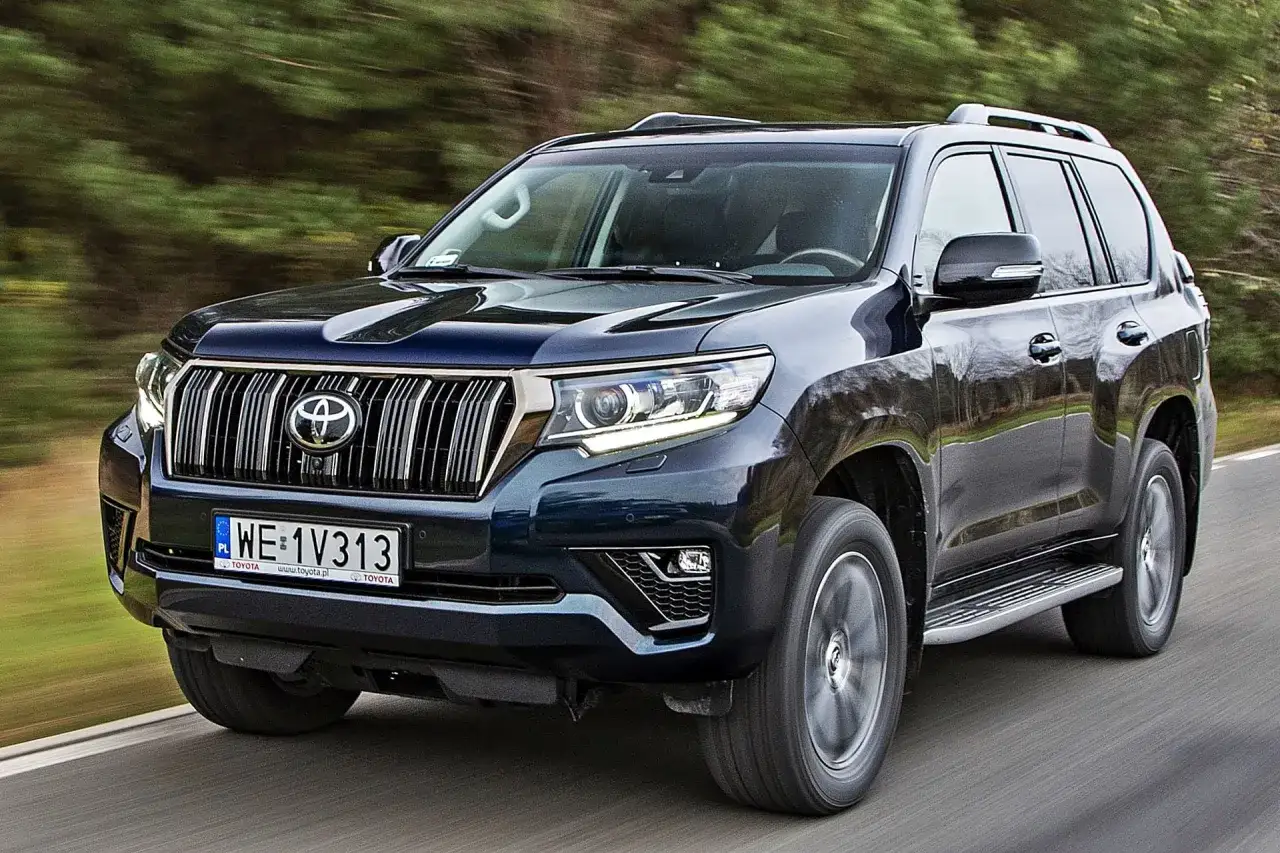 Ciemnogranatowy Toyota Land Cruiser 2024 pędzi po drodze, otoczony zielenią.