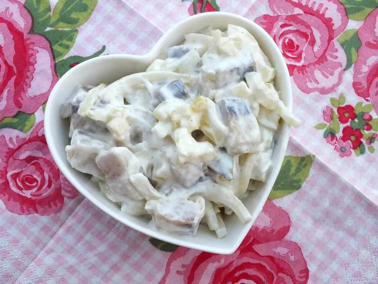 Pyszny przepis na śledzie w śmietanie z jabłkiem i ogórkiem. Sałatka w kształcie serca, podana na różowej, kwiecistej serwetce.