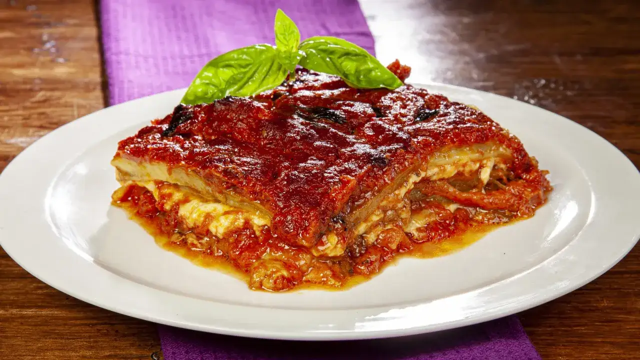 Una fetta di parmigiana di melanzane ricetta originale, con pomodoro e basilico fresco, servita su un piatto bianco.