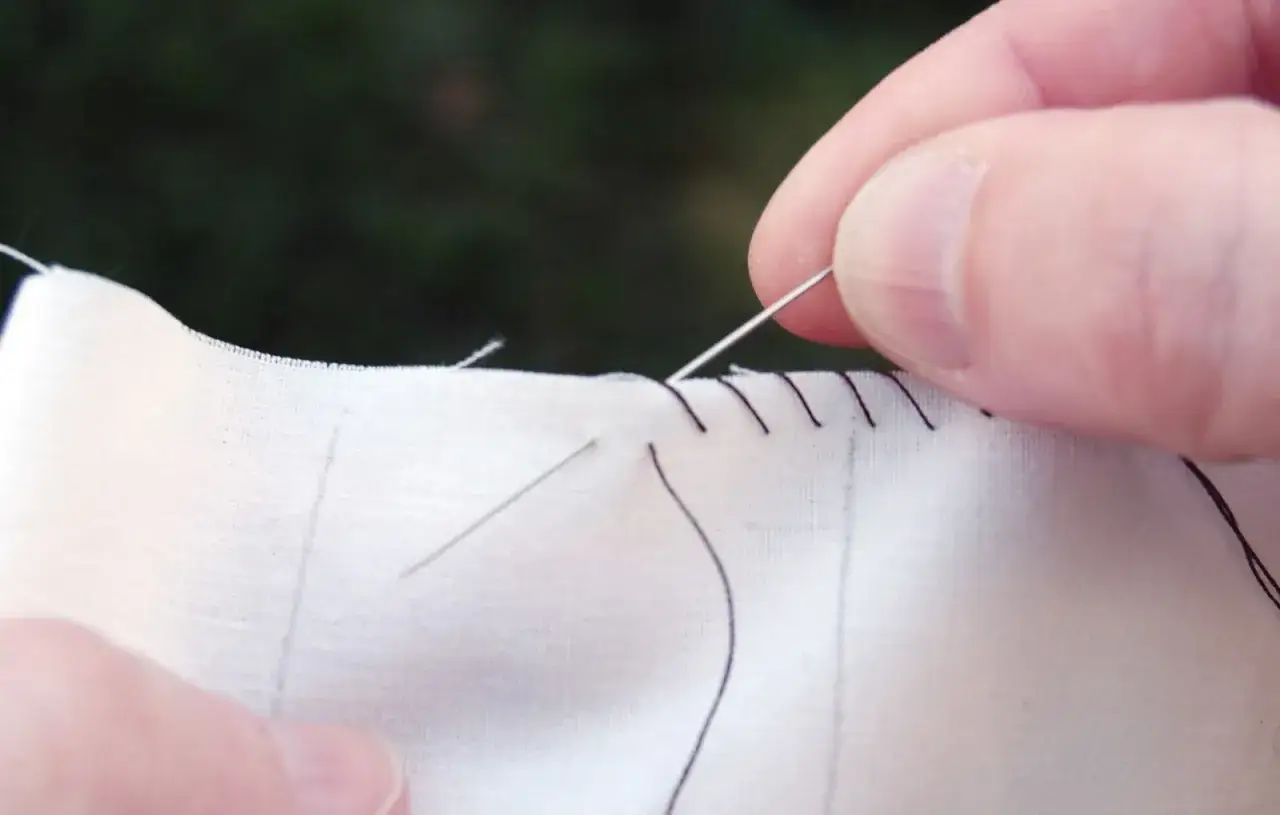 Close-up of a hand demonstrating how to do an invisible stitch on white fabric with dark thread.