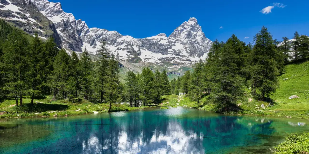 Lago alpino cristallino con montagne innevate e boschi di conifere. Un luogo incantevole per ammirare la natura e, con un po' di fortuna, avvistare un camoscio.