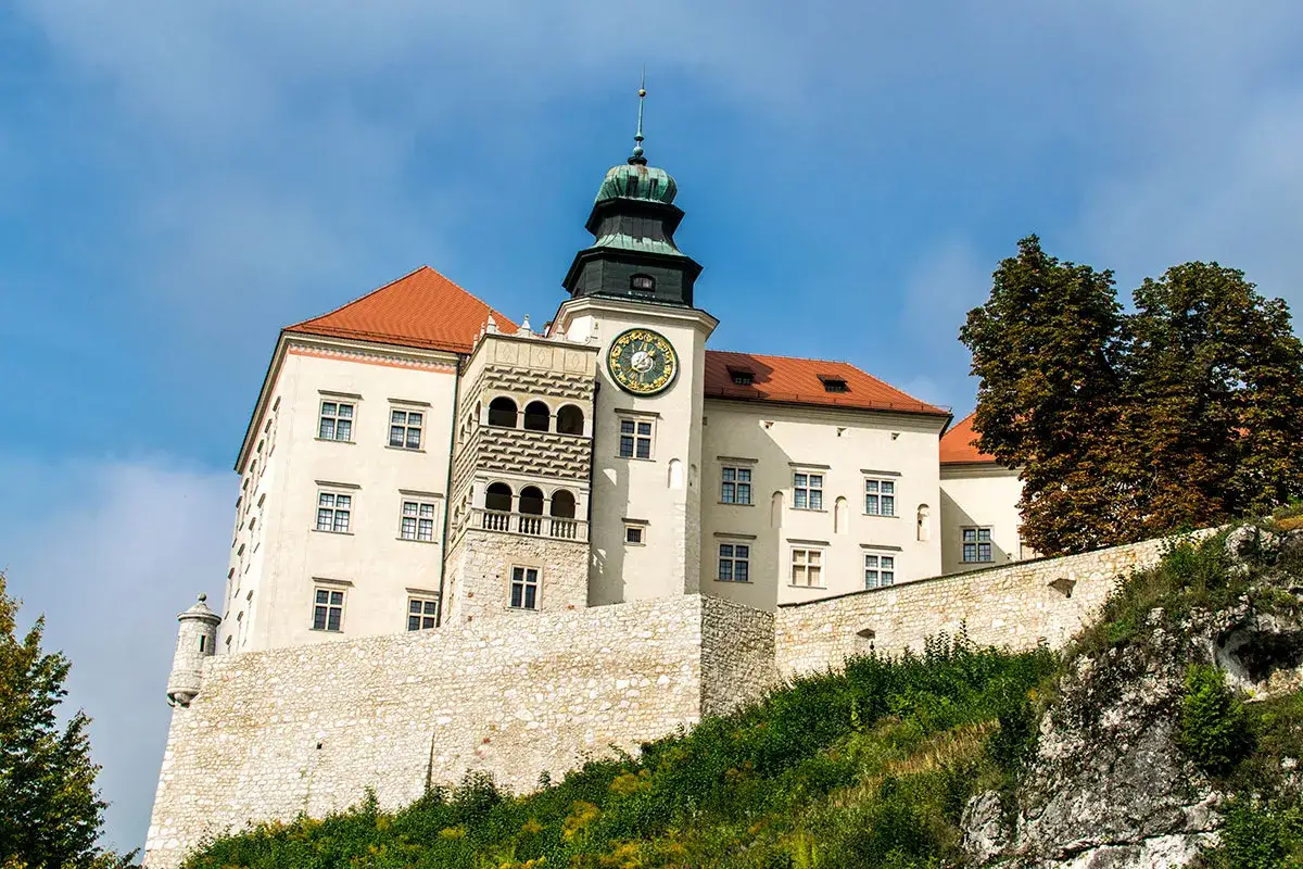 Zamek w Pieskowej Skale, majestatyczna budowla na skale, z wieżą zegarową i czerwonym dachem, otoczony zielenią.