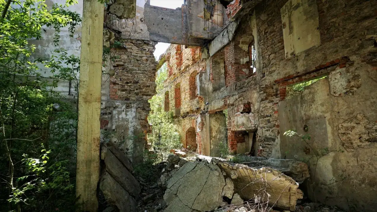 Ruiny budynku, zarośnięte zielenią, z widocznymi ceglanymi ścianami i gruzem. Idealne urbex miejsca dla poszukiwaczy przyg&oacute;d.