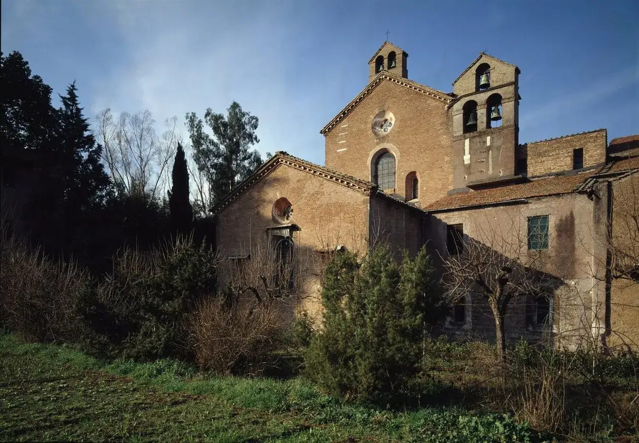 A brick church with bell towers stands against a blue sky. In the foreground, bare branches and evergreen bushes surround the building, hinting at the presence of the three fountains nearby.