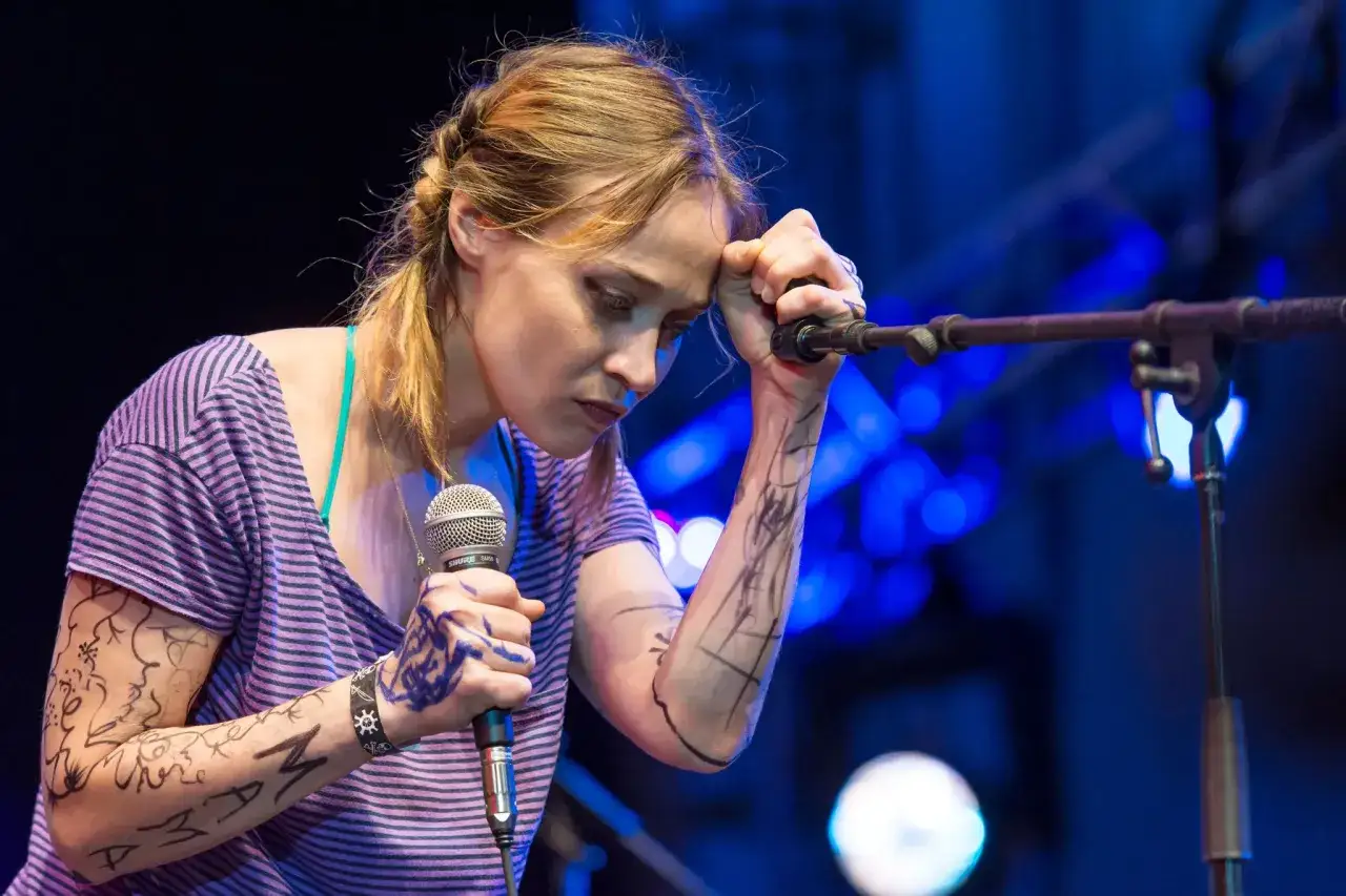 Fiona Apple, les bras couverts de dessins, chante intensément dans un micro, le front appuyé sur sa main.