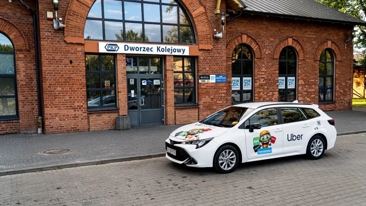 Biały samoch&oacute;d Ubera z naklejkami, zaparkowany przed budynkiem dworca kolejowego. Kierowca z własnym samochodem czeka na pasażer&oacute;w.