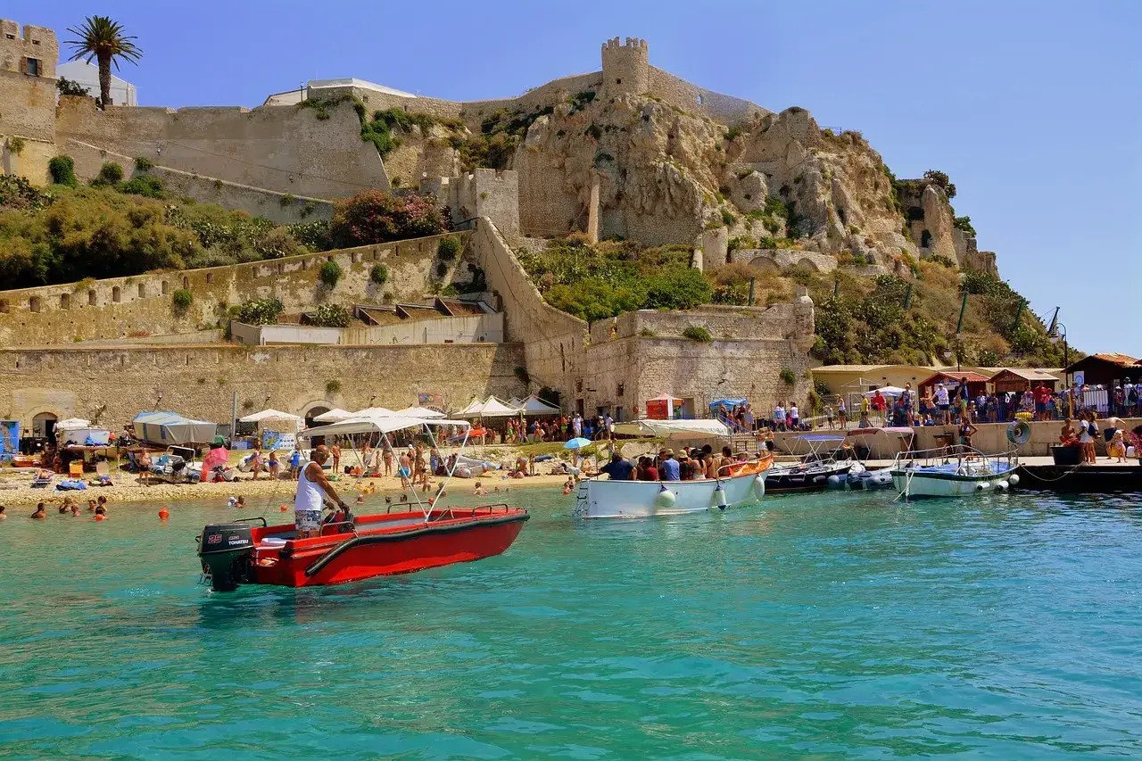 Un porto vivace con barche e gente sulla spiaggia, sotto un'antica fortezza che domina le isole della Puglia.