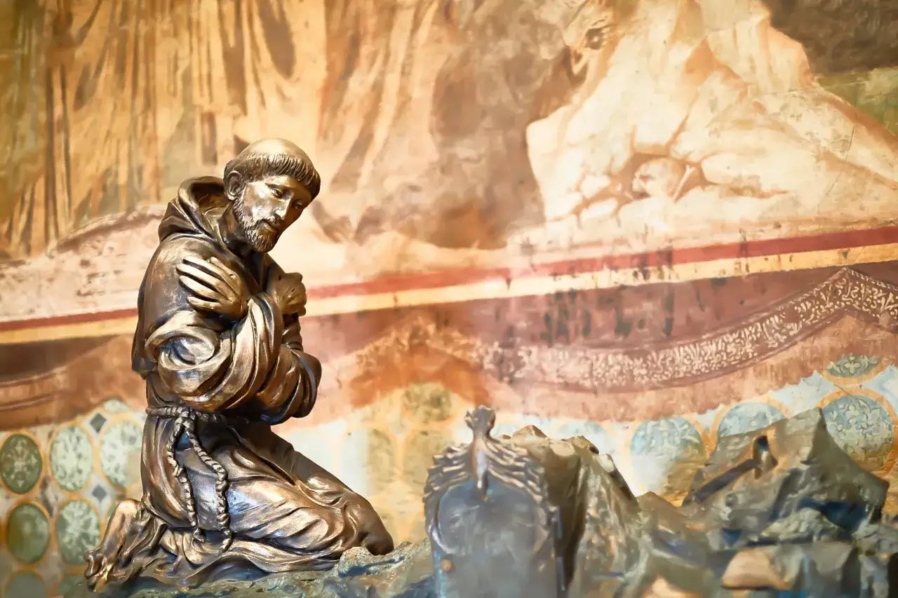 Bronzestatue von **St. Franziskus** kniend im Gebet vor einer Wandmalerei.