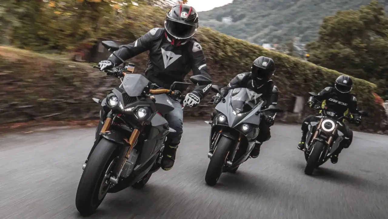 Tres pilotos en motos energicas recorren una carretera sinuosa entre &aacute;rboles.
