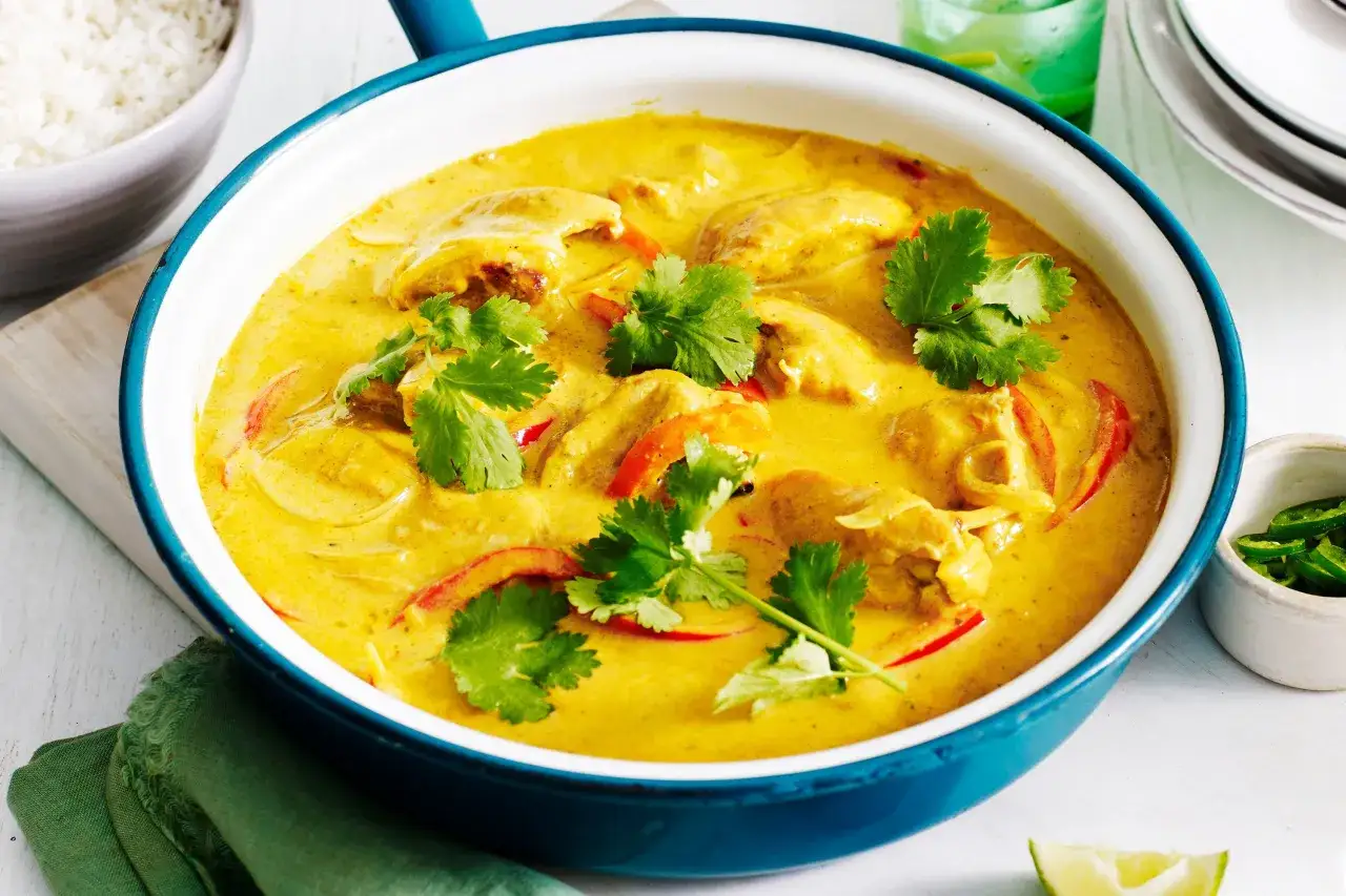 Aksamitny kurczak z mango po tajsku w żółtym sosie, ozdobiony kolendrą i papryką. Obok miska ryżu i zielone chili.