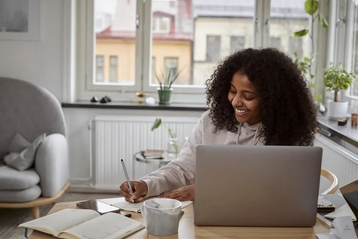 Glad kvinna pluggar frist&aring;ende kurser distans vid sin laptop. Hon skriver anteckningar i en bok.