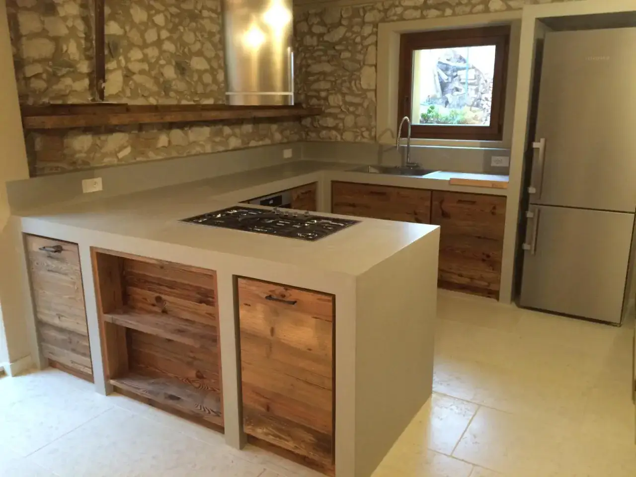 Progetto cucina in muratura moderna con isola centrale, piano in cemento, finiture in legno grezzo e cappa in acciaio.