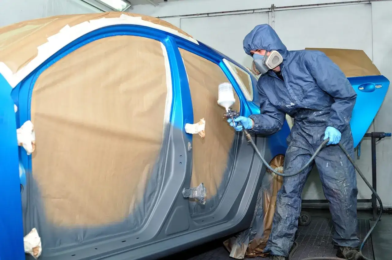Carrozziere in tuta spruzza vernice su un'auto blu. Informazioni su quanto costa cambiare il colore della macchina sul libretto.