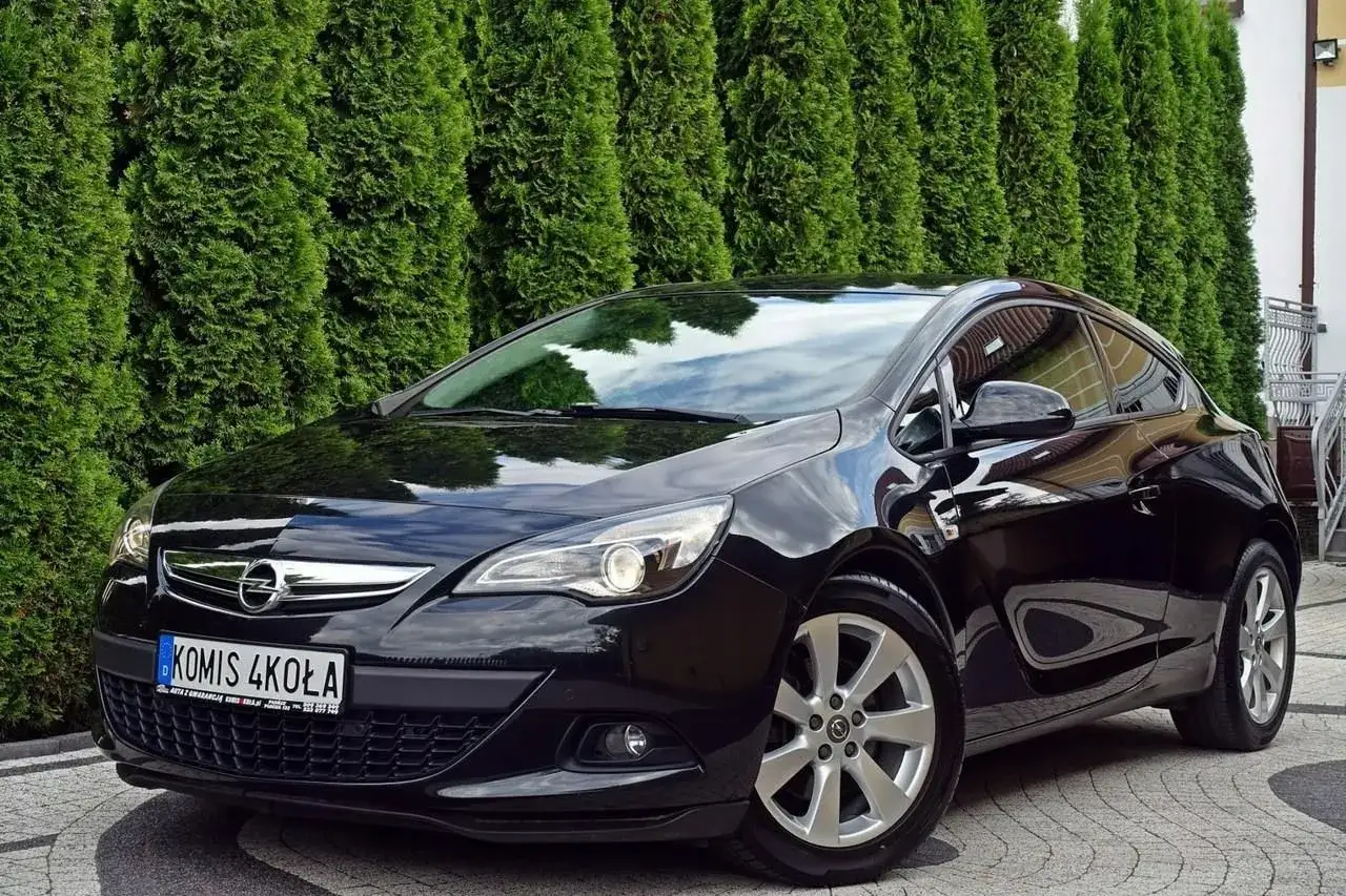 Błyszczący czarny Opel Astra GTC z przyciemnianymi szybami, zaparkowany przed żywopłotem z tuj.