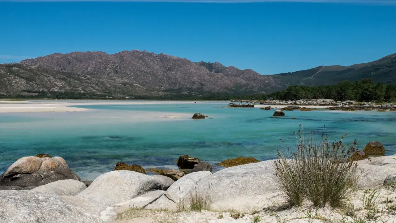 Aguas turquesas y arena blanca en la playa de San Pedro, Galicia, con montañas rocosas y pinos de fondo.