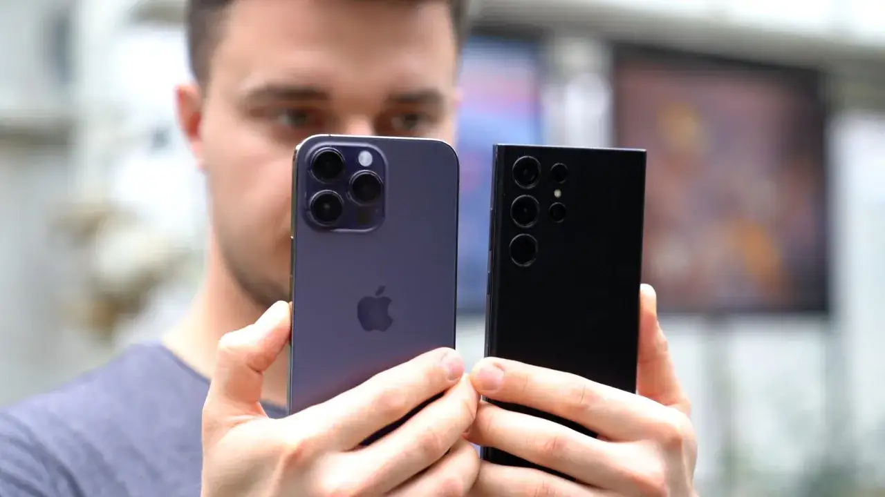 Mężczyzna trzyma dwa smartfony, iPhone i Samsung, porównując ich aparaty. Oba telefony mają dobry aparat, idealny do robienia zdjęć.