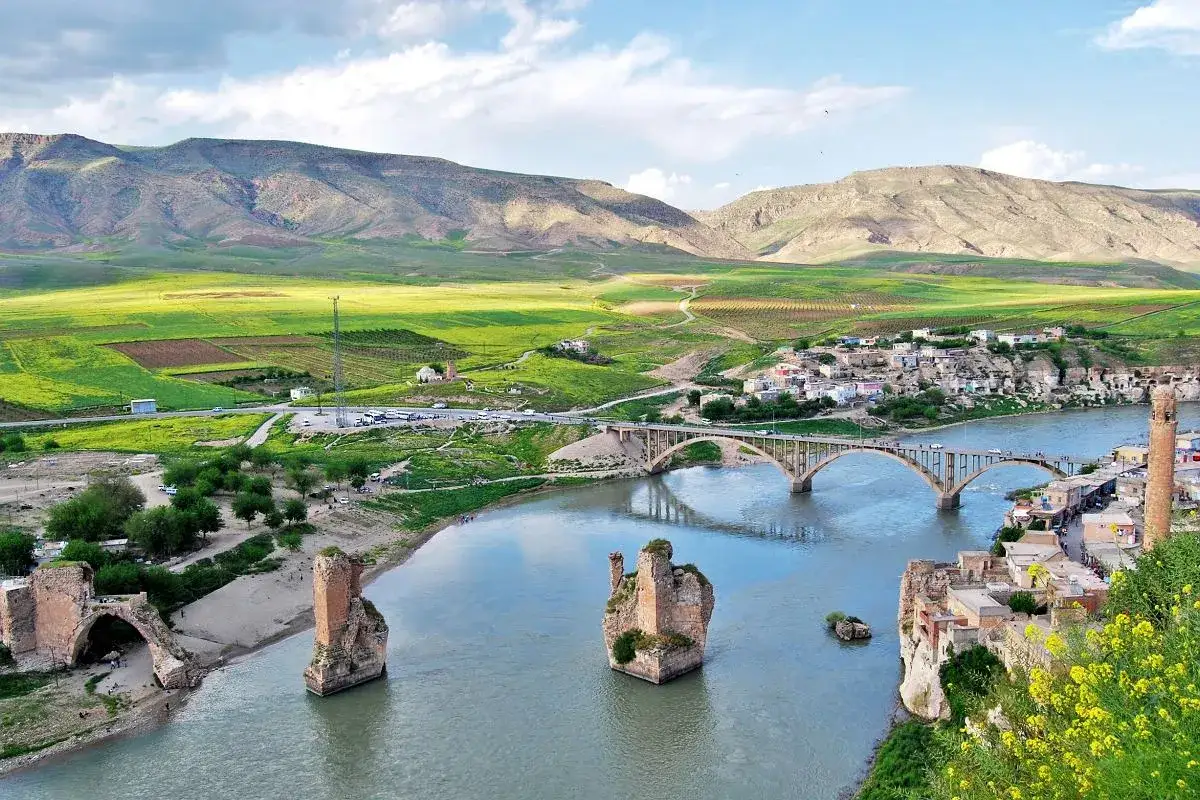 Malowniczy widok na rzekę i most w Hasankeyf, otoczony zielonymi wzgórzami i historycznymi ruinami.
