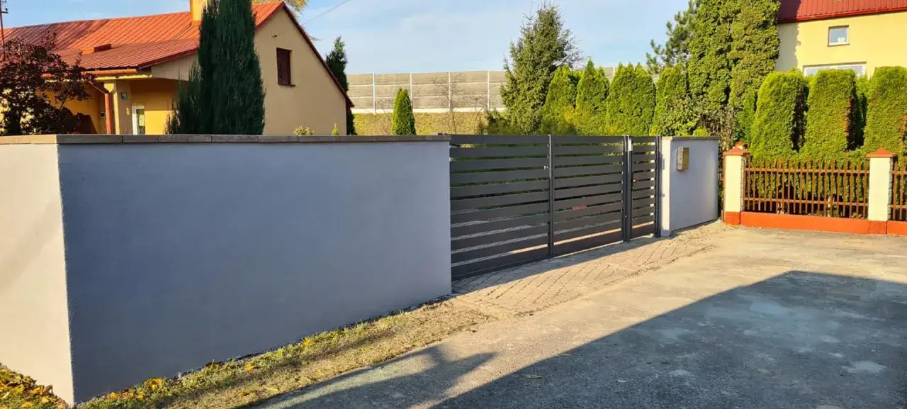 Nowoczesne ogrodzenie z szarego, tynkowanego muru i metalowej bramy.