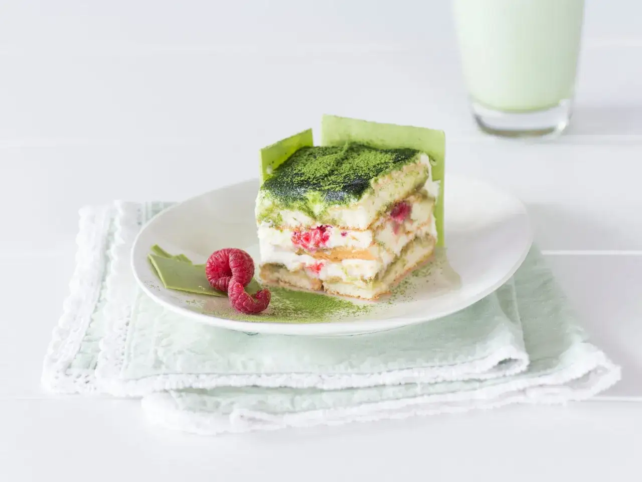 Ein Stück Matcha Torte mit Himbeeren und grüner Schokolade, bestäubt mit Matcha-Pulver.